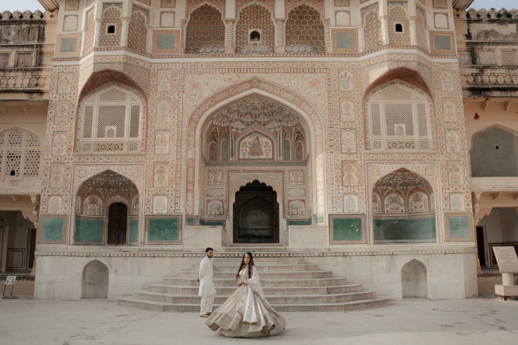 A couple poses in front of the highly detailed Ganesh Pol gateway during an Amer Fort pre-wedding photoshoot, with the bride twirling in a white lehenga against the backdrop of ancient frescoes and arched doorways.