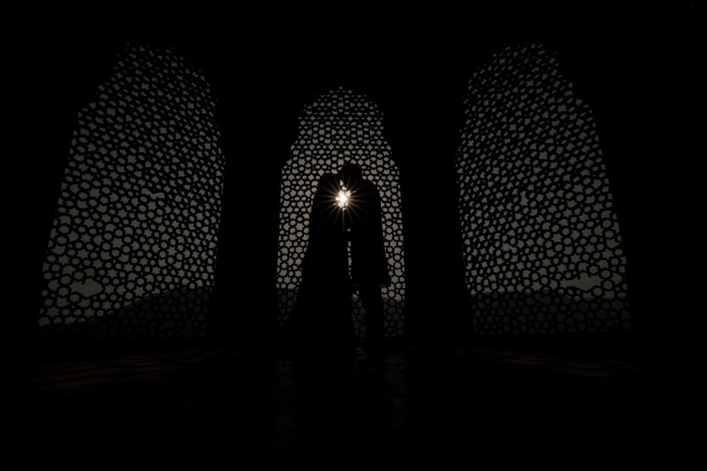 A silhouette of a couple embracing during an Amer Fort pre-wedding photoshoot, framed by three ornate Mughal-style arched windows with intricate geometric stone carvings (jali) as the sun glows between them.