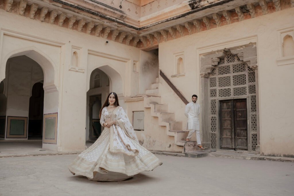 A wide-angle shot of a couple during an Amer Fort pre-wedding photoshoot standing before the magnificent Ganesh Pol gateway. The bride is twirling in a voluminous white and gold lehenga while the groom stands behind her, framed by the gate's famous frescoes and mosaic art.