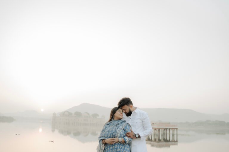 A romantic couple posing for a Jal Mahal pre-wedding photoshoot in Jaipur with the water palace and a soft sunrise in the background.
